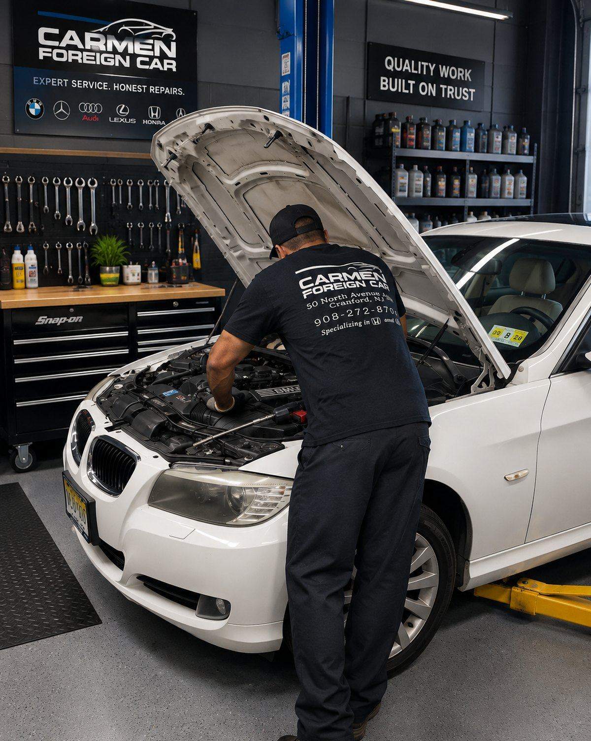 Technician in a Carmen's Foreign Car shirt working under the hood of a white sedan in the Cranford shop